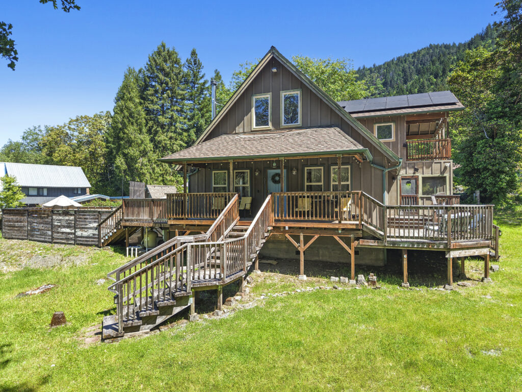 A brown wooden house with a wrap-around front porch and solar panels on the roof, surrounded by lush green grass and trees on a sunny day.