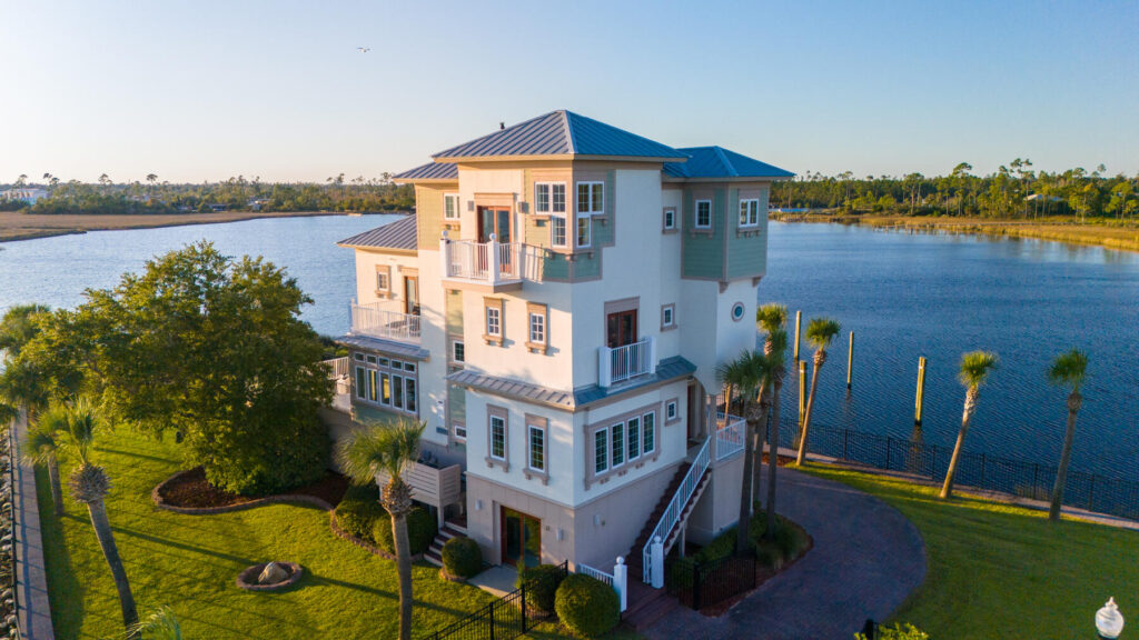 A three-story, modern coastal waterside home with balconies sits beside a calm river or lake, surrounded by green grass, palm trees, and a bright blue sky during sunrise or sunset.