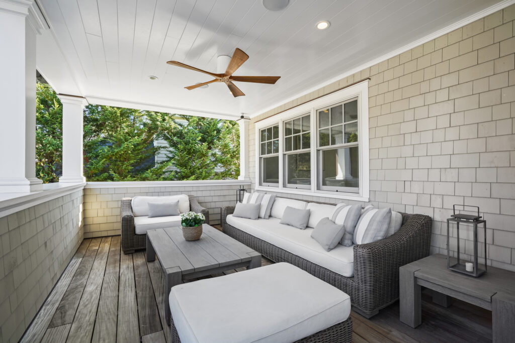 A cozy front porch with wicker furniture, including a sofa, chairs, and ottomans with white cushions, a wooden coffee table, ceiling fan, lantern, and potted plant—surrounded by shingle siding and windows.