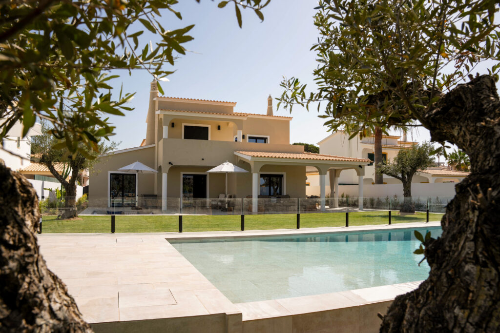 Two leafy trees frame the view of a modern beige house with a tiled roof, large windows, and a spacious patio. A backyard pool sparkles beside a manicured lawn under a clear sky, perfect for relaxing during gemini season.