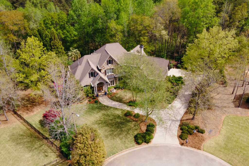 A large two-story house with a brown roof sits at the end of a curved driveway, surrounded by well-kept landscaping and green trees, viewed from above on a sunny Gemini season day.