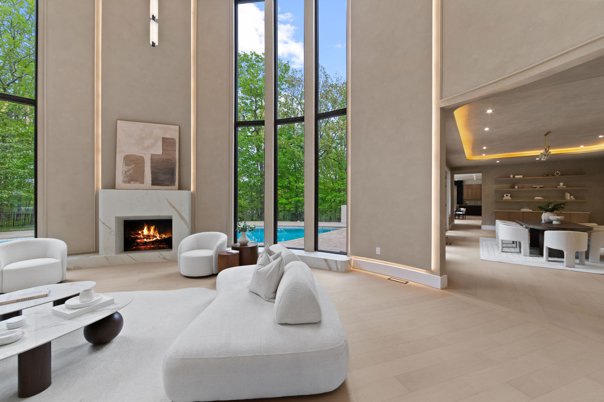 IMG_1232 A modern living room with high ceilings, tall windows showing greenery and a pool, light-colored walls and furniture, a lit fireplace, and an open view into a dining area—perfect for hosting an A-list June 2026 gathering.
