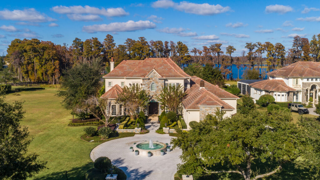 A large, elegant house with a red-tiled roof sits amid manicured lawns and trees. During gemini season, the circular driveway with a central fountain gleams, while a lake and lush trees complete the scene beneath a blue, partly cloudy sky.