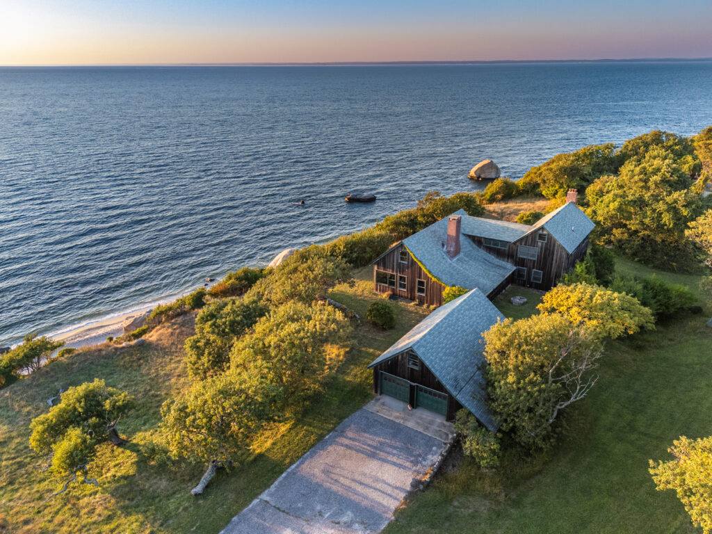 Aerial view of a house with a sloped roof nestled among trees on a grassy bluff, overlooking a calm ocean at sunset during gemini season, with a sandy beach and gentle waves below.