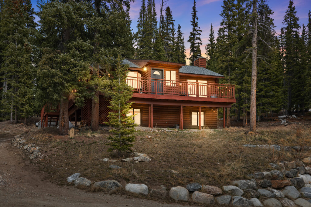 A cozy wooden cabin with glowing lights sits among tall pine trees at dusk, featuring a wraparound balcony and surrounded by a rocky, wooded landscape—perfect for unwinding during gemini season.