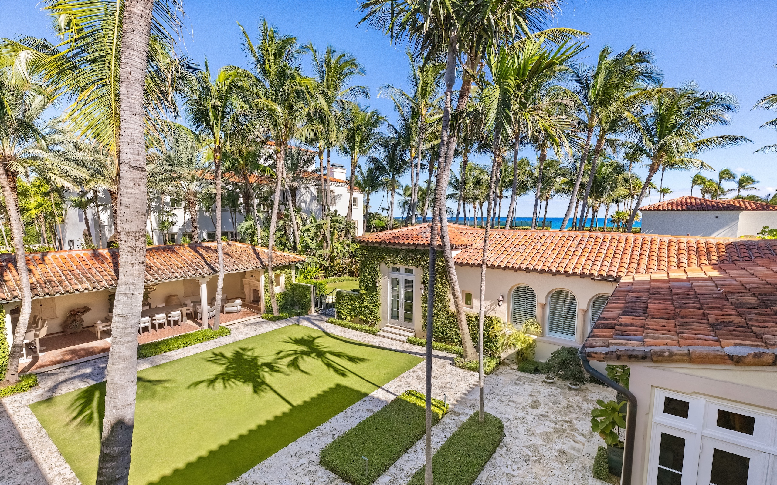 A Mediterranean-style villa courtyard at 115 Via La Selva features red-tiled roofs, palm trees, manicured hedges, stone pathways, and a green lawn, set near the ocean under a clear blue sky.