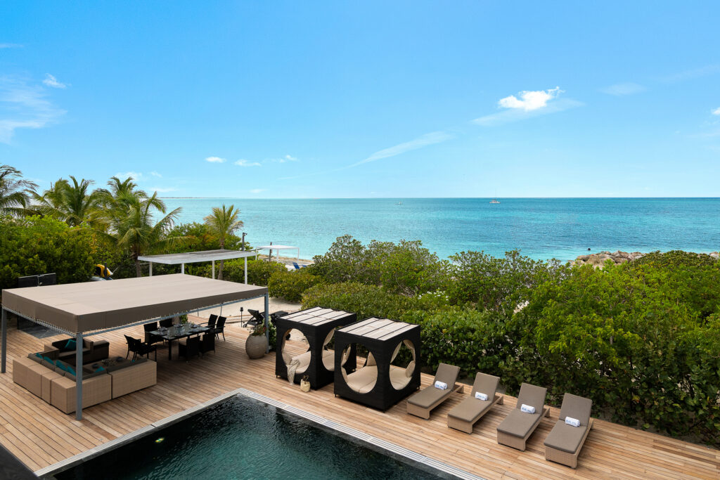 A luxurious beachfront deck with a shaded lounge area, sunbeds, cabanas, and a swimming pool overlooks clear blue ocean water under a bright sky, surrounded by lush greenery, palm trees, and elegant waterside homes.