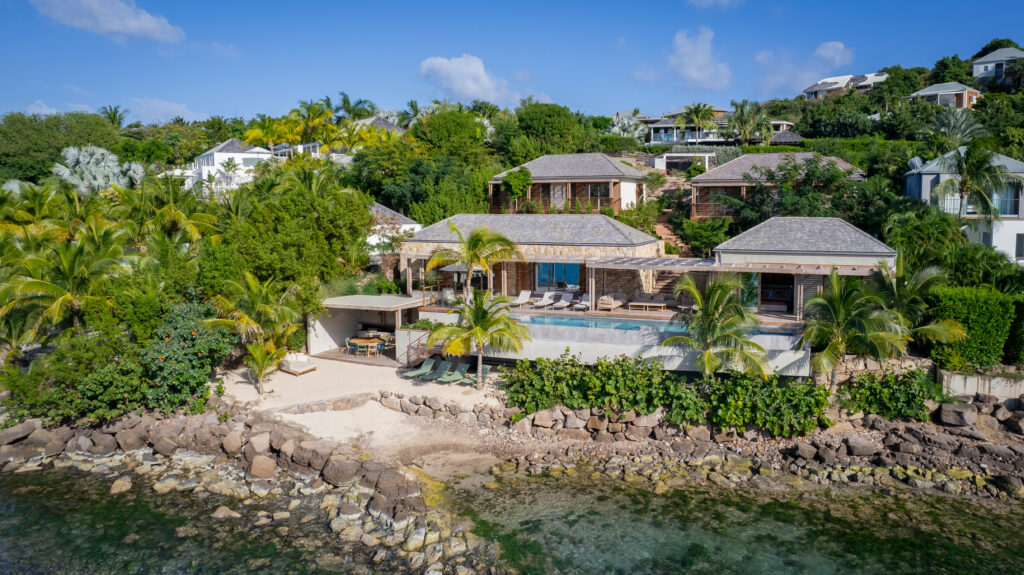 A luxurious waterside home with a pool and multiple buildings sits on a lush, tropical hillside overlooking clear, rocky shoreline and turquoise water under a blue sky with scattered clouds.
