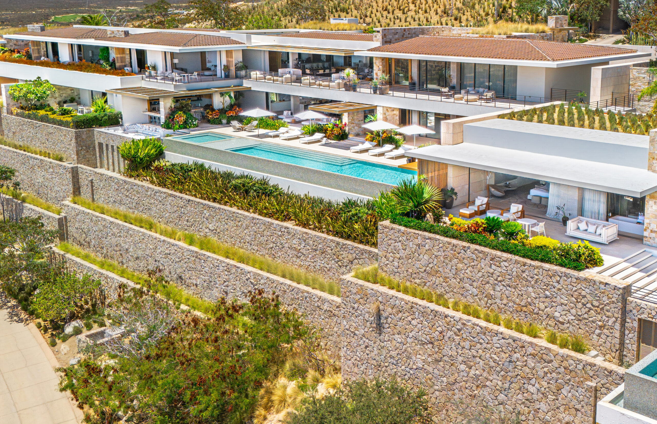 cabo-full A modern luxury villa with multiple terraces, an infinity pool, outdoor seating areas, and lush landscaping, surrounded by stone walls and set on a hillside—perfect for an A-list June 2026 getaway.