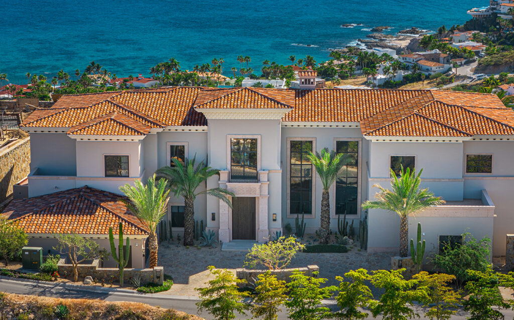 A large, luxurious villa with a red-tiled roof and palm trees in front, this stunning waterside home overlooks the ocean and coastal landscape under a clear, sunny sky.
