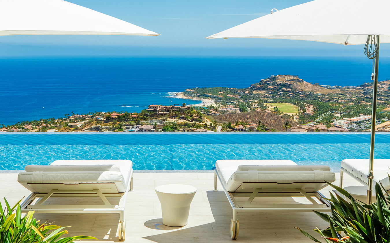 Two white lounge chairs and umbrellas face an infinity pool overlooking a coastline, with blue ocean, hills, and a clear sky in the background—a-list: June 2026. Lush greenery frames the foreground.