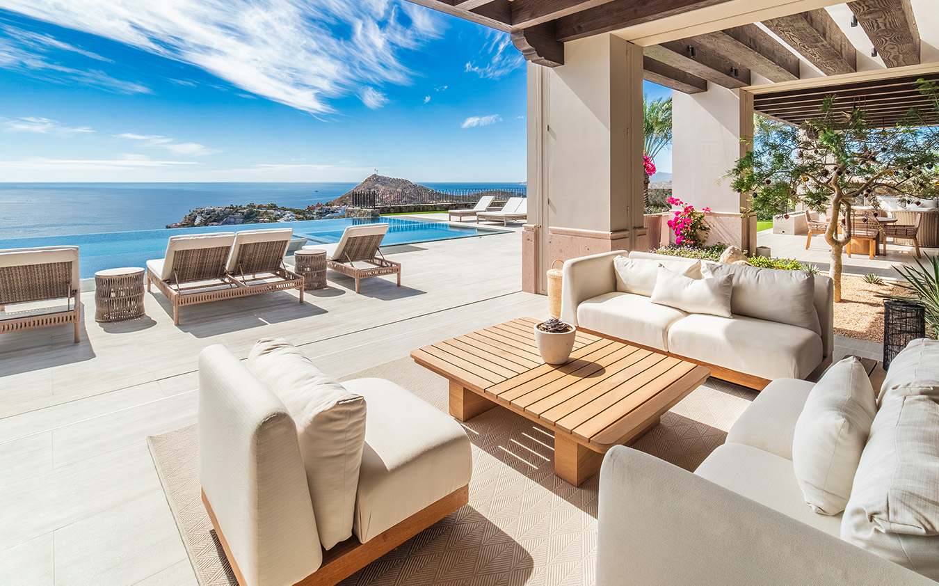 Modern outdoor patio with white cushioned furniture, wooden table, and lounge chairs overlooking a pool and scenic ocean view—a perfect retreat for waterside homes under a bright blue sky with scattered clouds.