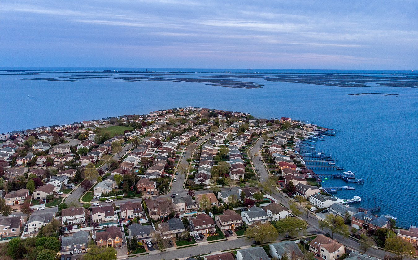 Aerial view of a coastal suburban neighborhood with rows of houses, docks, and boats along the shoreline, bordered by a large body of water under a cloudy sky.
