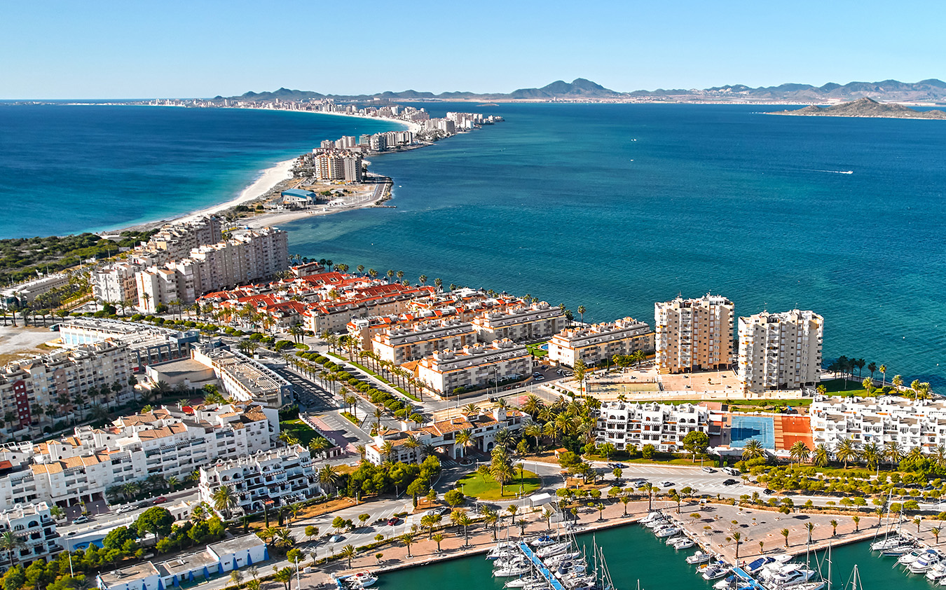 Aerial view of a coastal city with high-rise buildings, marinas with boats, and a narrow strip of land stretching into the blue sea, bordered by sandy beaches on both sides and distant mountains on the horizon.