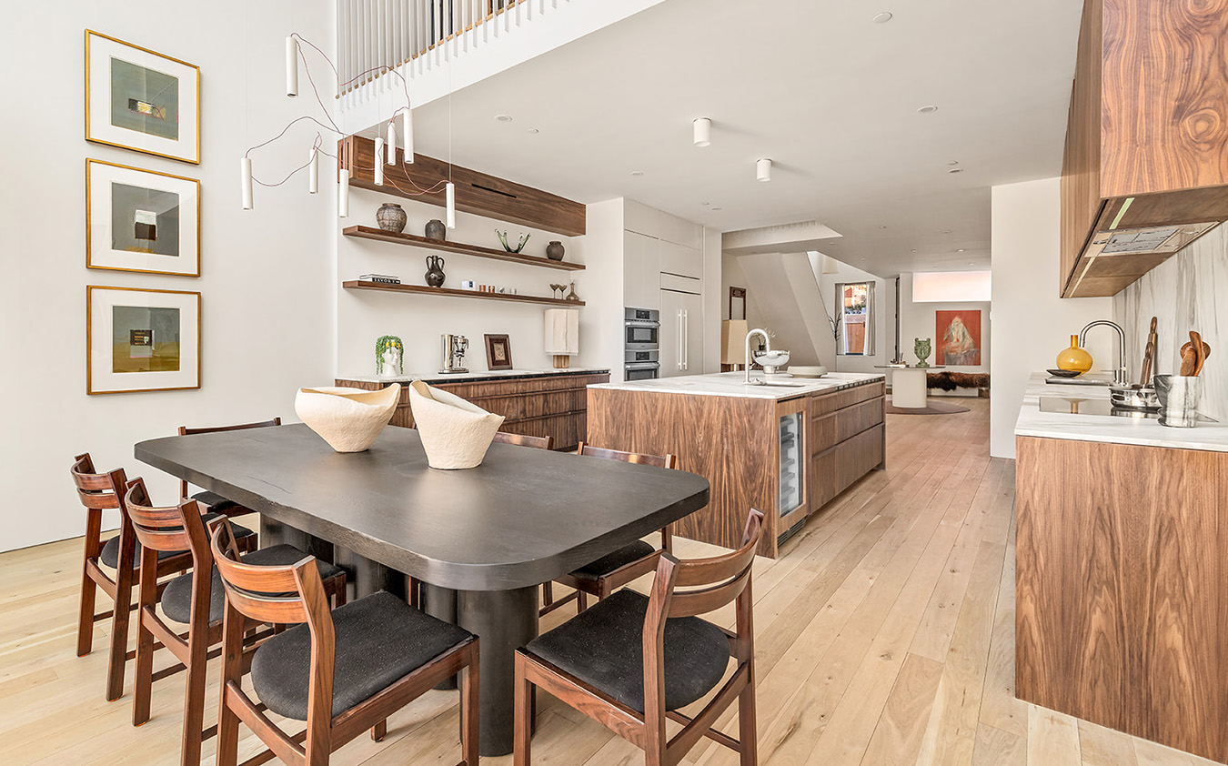 Modern open kitchen and dining area with wood cabinetry, a black dining table with six chairs, white walls, open shelves with ceramics, and light wood flooring. Natural light brightens the space.