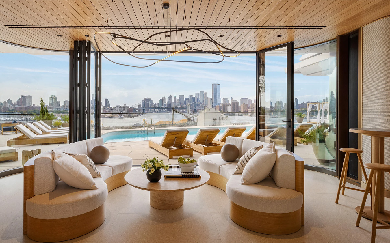 Modern living room with round sofas and a circular coffee table, opening to a patio with lounge chairs by a pool, overlooking a city skyline across the water on a sunny day.