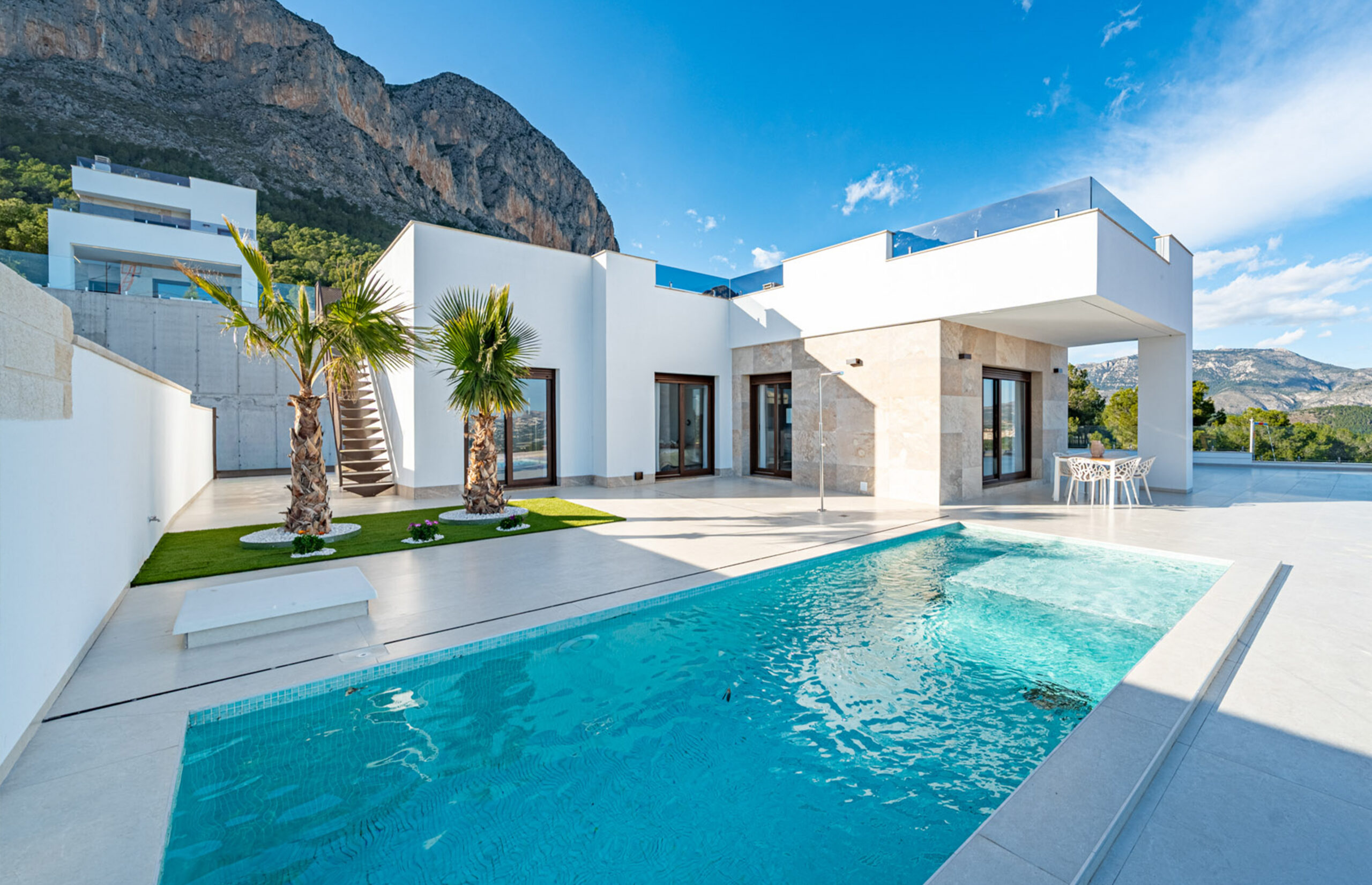 spain Modern white villa with large windows, palm trees, and a rectangular swimming pool, set against a mountainous backdrop under a clear blue sky. Outdoor seating and minimalist landscaping evoke an a-list: June 2026 retreat.