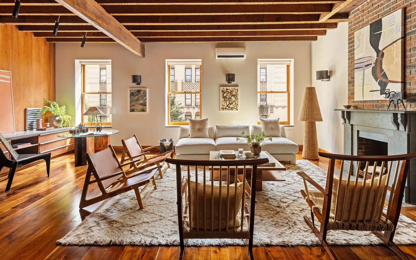 A stylish living room with exposed wooden beams, large windows, a white sofa, wooden chairs, a cozy rug, and modern decor. A brick fireplace and artwork add warmth to the space, which features abundant natural light.