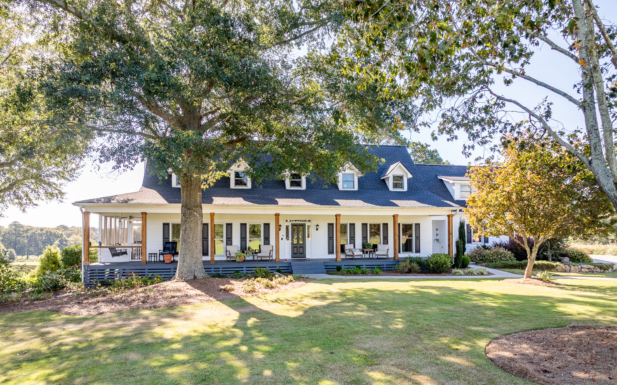 A white two-story house with a dark roof features a spacious front porch that wraps around, set on a green lawn surrounded by trees.