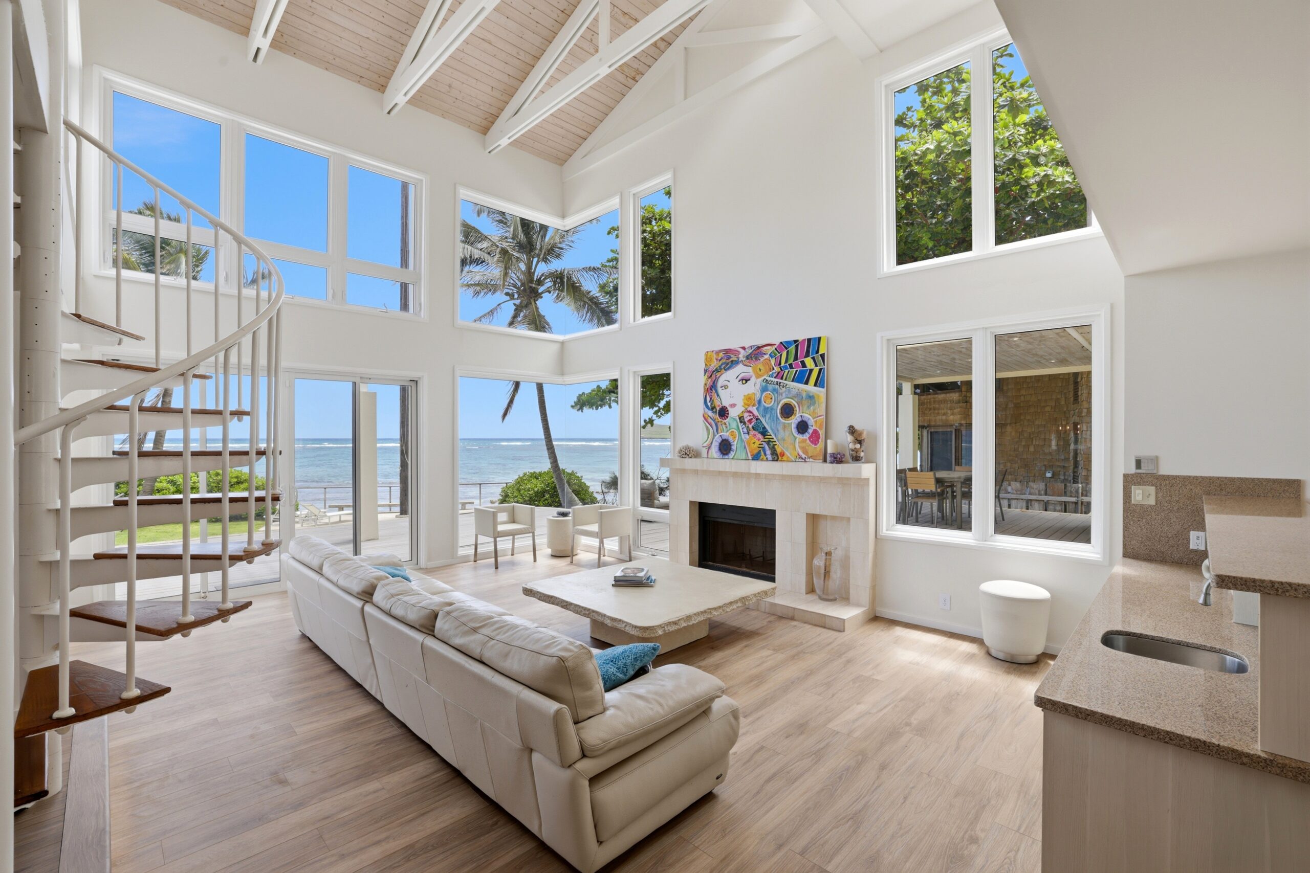 Spacious, modern living room with high ceilings, large windows with ocean views, a white spiral staircase, light wood floors, a white sofa, a fireplace, colorful artwork, and abundant natural light—a true A-list retreat for June 2026.