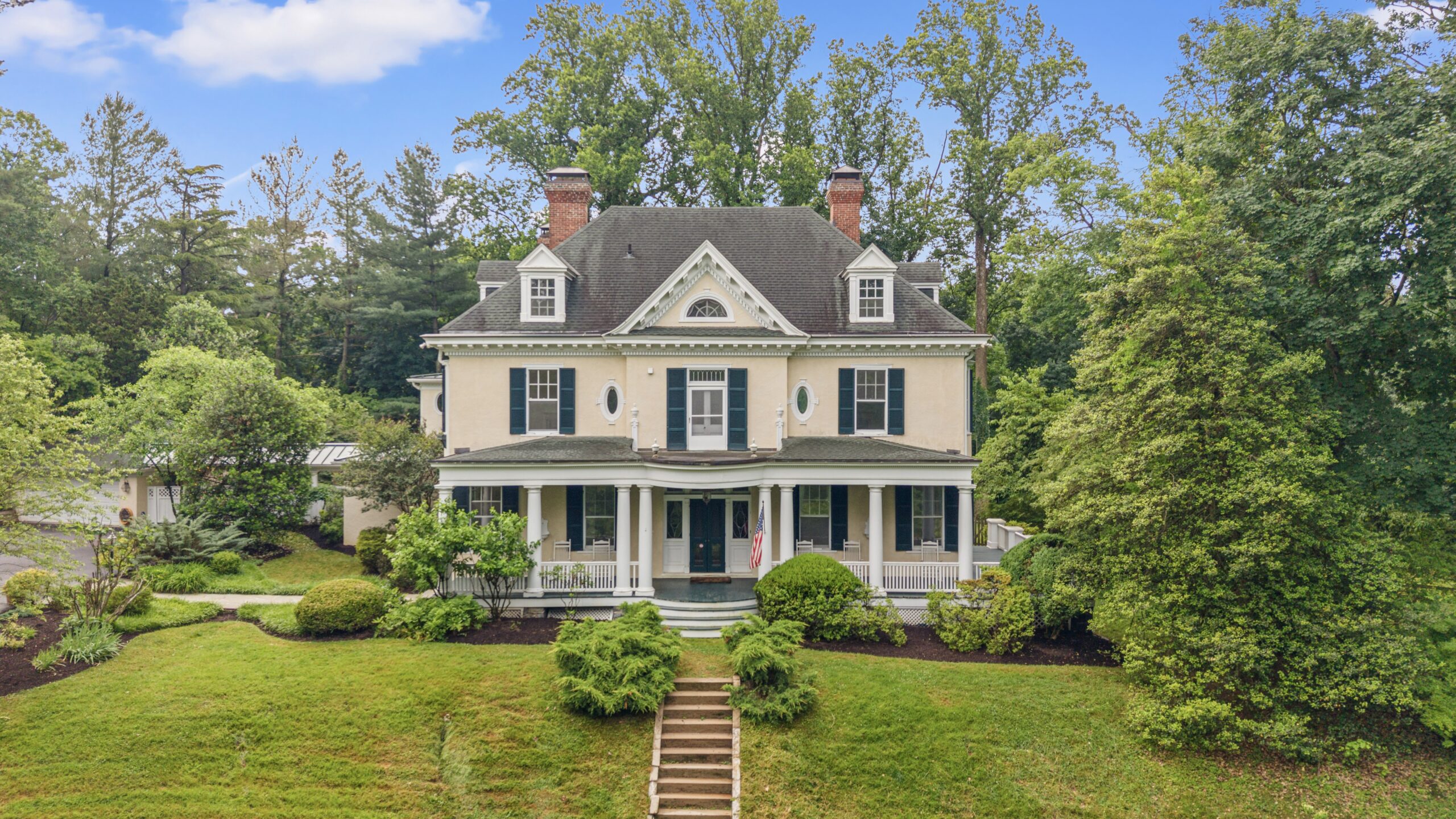 A large, two-story historic house with beige siding, dark shutters, and a wraparound porch, surrounded by green trees and landscaping under a blue June 2026 sky with scattered clouds—a true A-list gem.