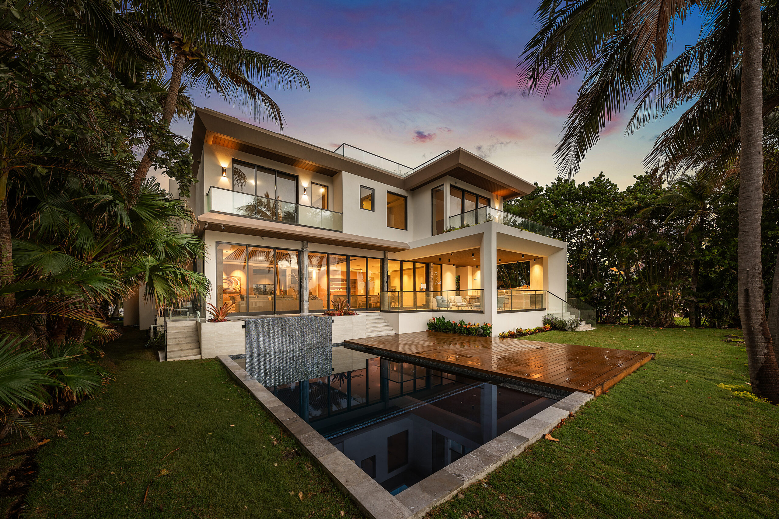 Modern two-story house with large windows, a wooden deck, small pool with water feature, surrounded by lush greenery and palm trees at sunset. Warm interior lights create an inviting atmosphere—an A-list June 2026 oasis.