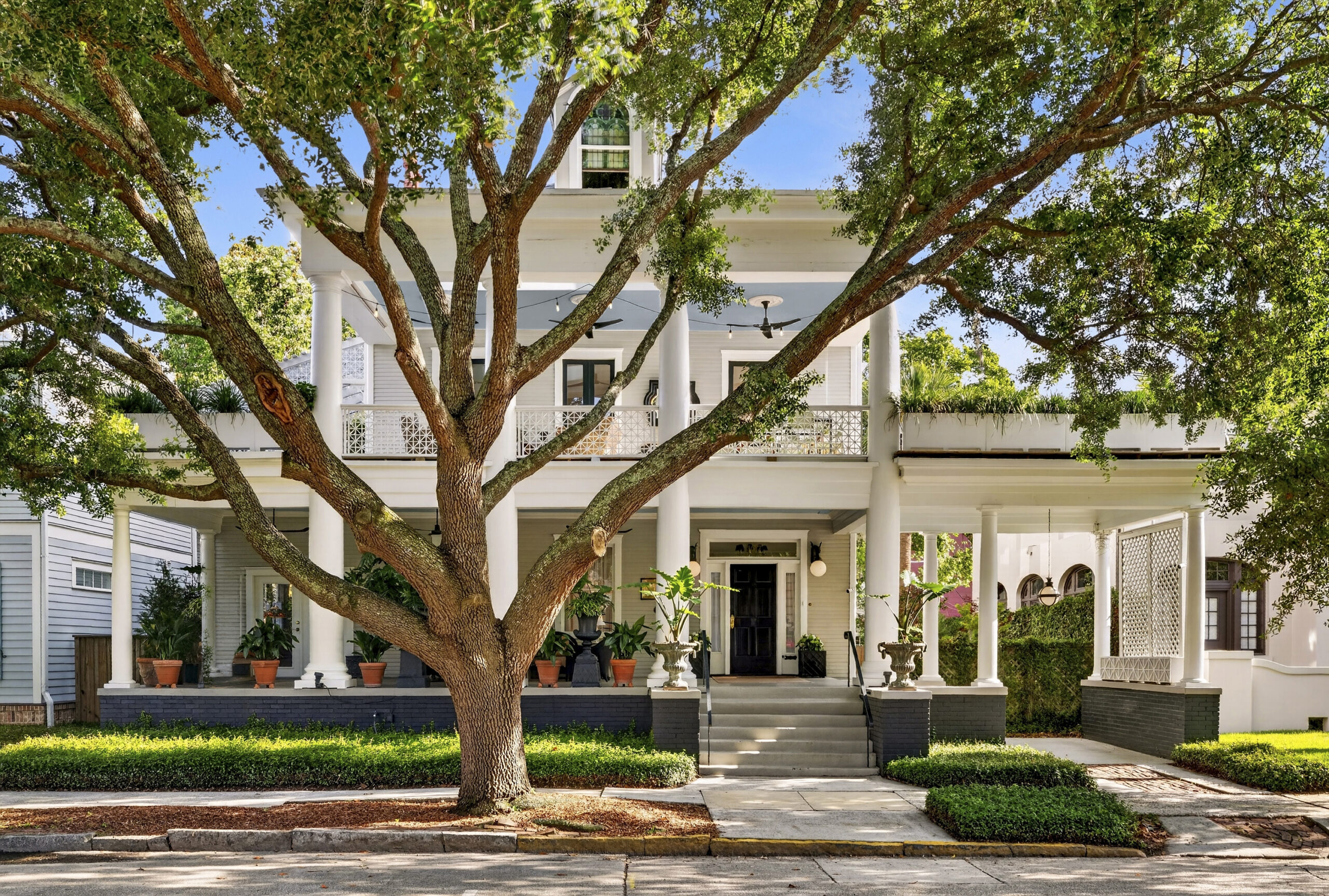Large, historic two-story house with white columns and wraparound porch, partially shaded by a broad oak; potted plants line the porch and steps lead up to the entrance—an A-list gem set for June 2026 charm.