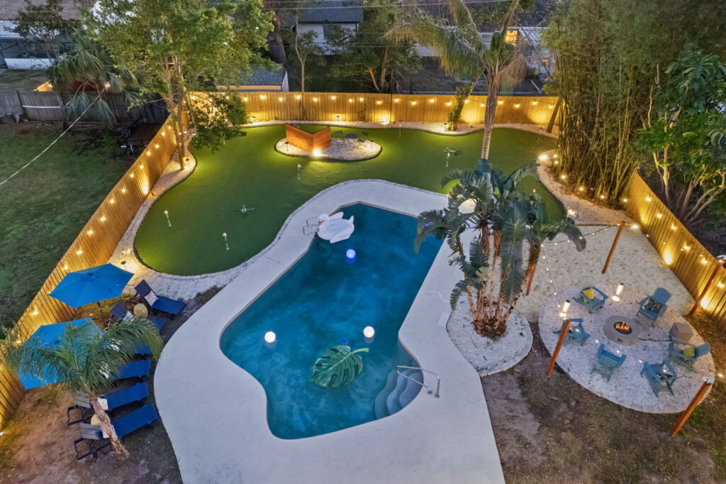 Aerial view of a backyard oasis featuring a pool with floating toys, lounge chairs, palm trees, string lights on a wooden fence, artificial turf, a cozy fire pit area with seating, and shaded blue umbrellas.