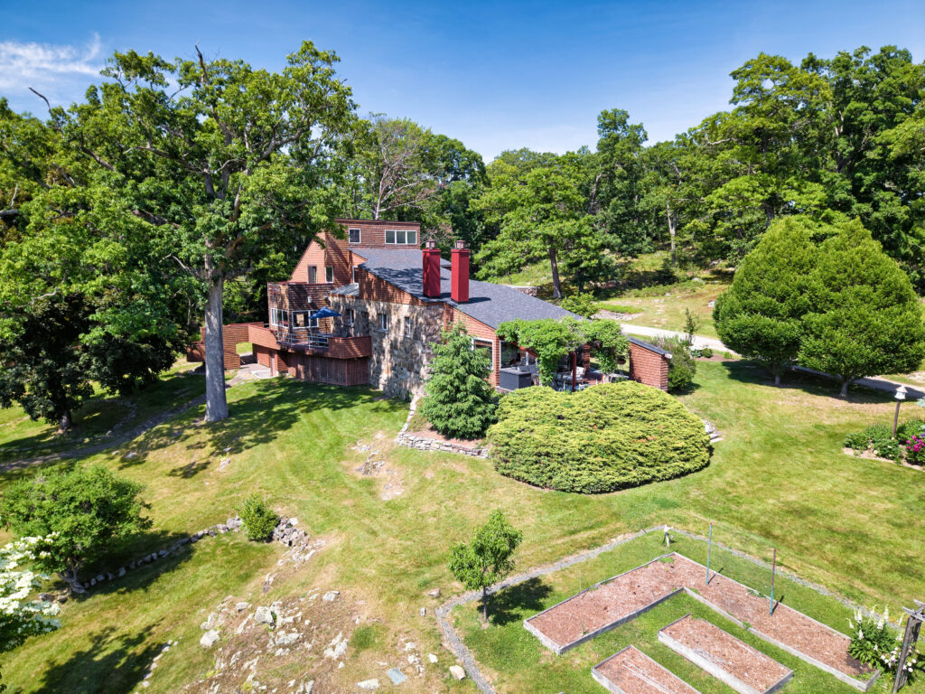 A large brick and stone house with a deck overlooks a spacious backyard, surrounded by lush green trees and expansive lawn, with a small garden area and clear blue sky overhead.