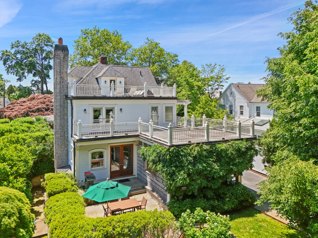 A two-story house with a large balcony and white railing overlooks a spacious backyard filled with lush greenery. A patio below features a table and green umbrella, while trees and neighboring houses sit beneath the blue sky.