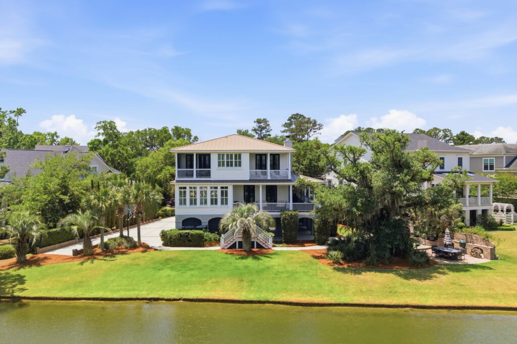 A two-story house with large windows, a porch, and a raised entrance sits by a calm pond, featuring a lush backyard surrounded by green grass, palm trees, and other houses under a bright blue sky.