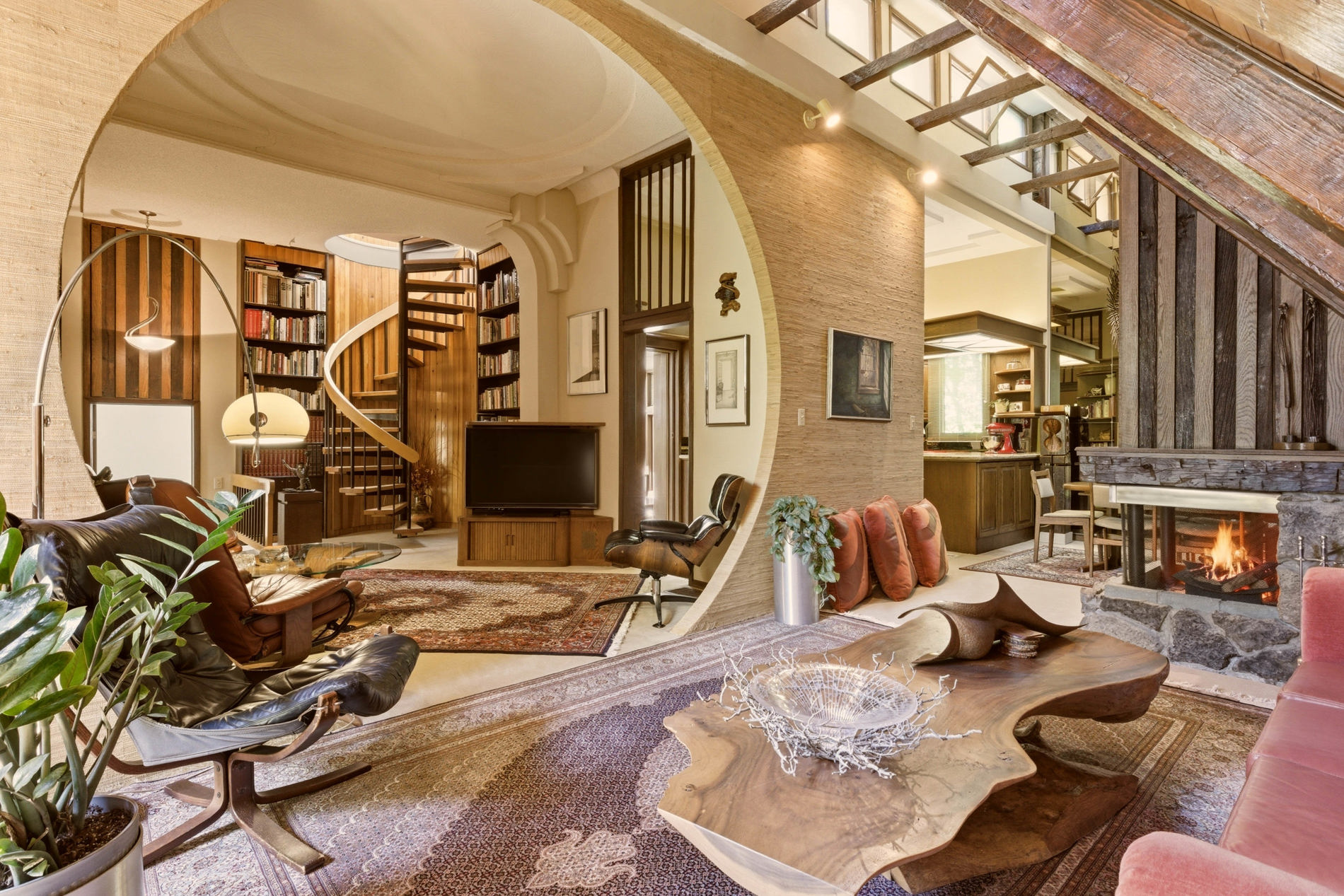 A spacious living room features a large circular opening in the wall, a spiral staircase, eclectic furniture, and a unique wood coffee table—delivering an inviting atmosphere that feels ready for an A-list June 2026 gathering.
