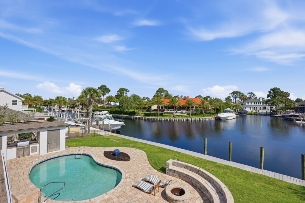 A stunning backyard oasis features a circular swimming pool, hot tub, lounge chairs, and a private dock with boats on a calm canal—all surrounded by palm trees and houses under a blue sky with scattered clouds.