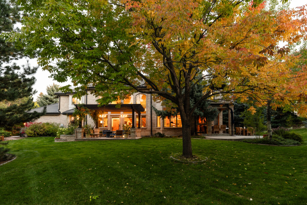 A large house with glowing windows sits behind a lush green lawn, a backyard tree with autumn-colored leaves creating a cozy and inviting atmosphere at dusk.