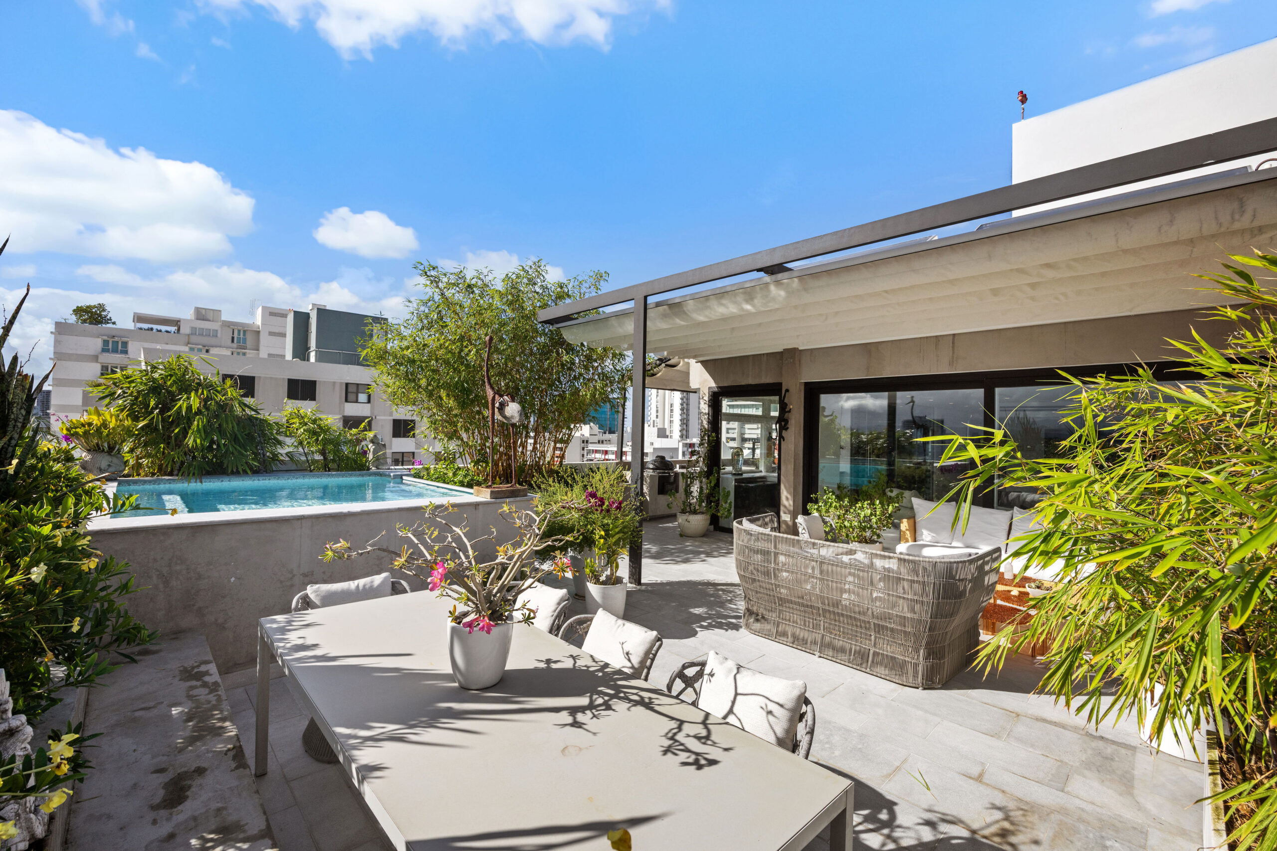 A modern outdoor patio with a dining table and chairs, lush green plants, and a swimming pool, perfect for hosting an A-list: June 2026 gathering. Large glass doors connect the space to the house under a bright blue sky with scattered clouds.