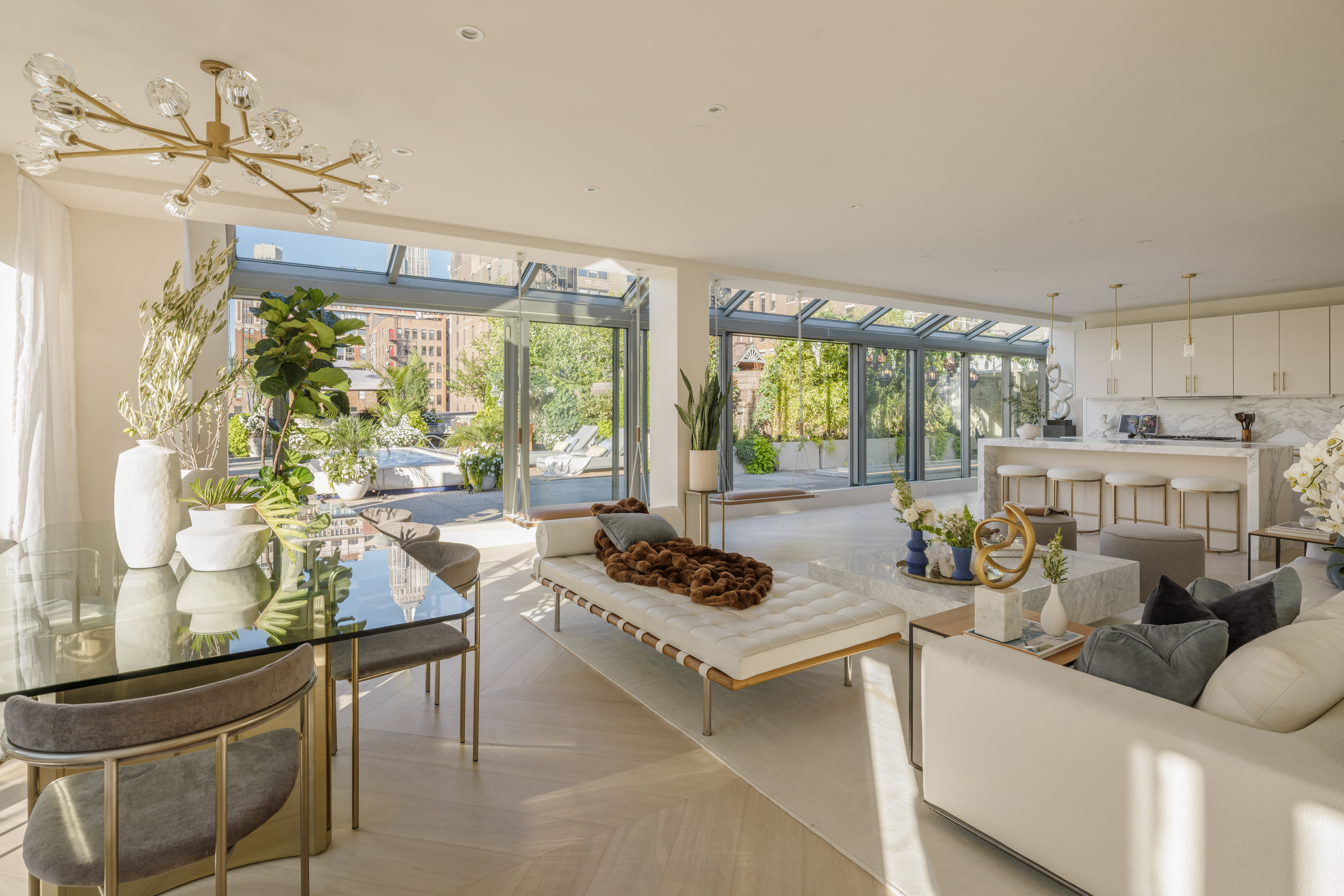 Sunlit luxury penthouse living space with open-concept dining and kitchen, featuring marble island, modern chandelier, floor-to-ceiling glass walls, and landscaped terrace views in a contemporary urban residence.
