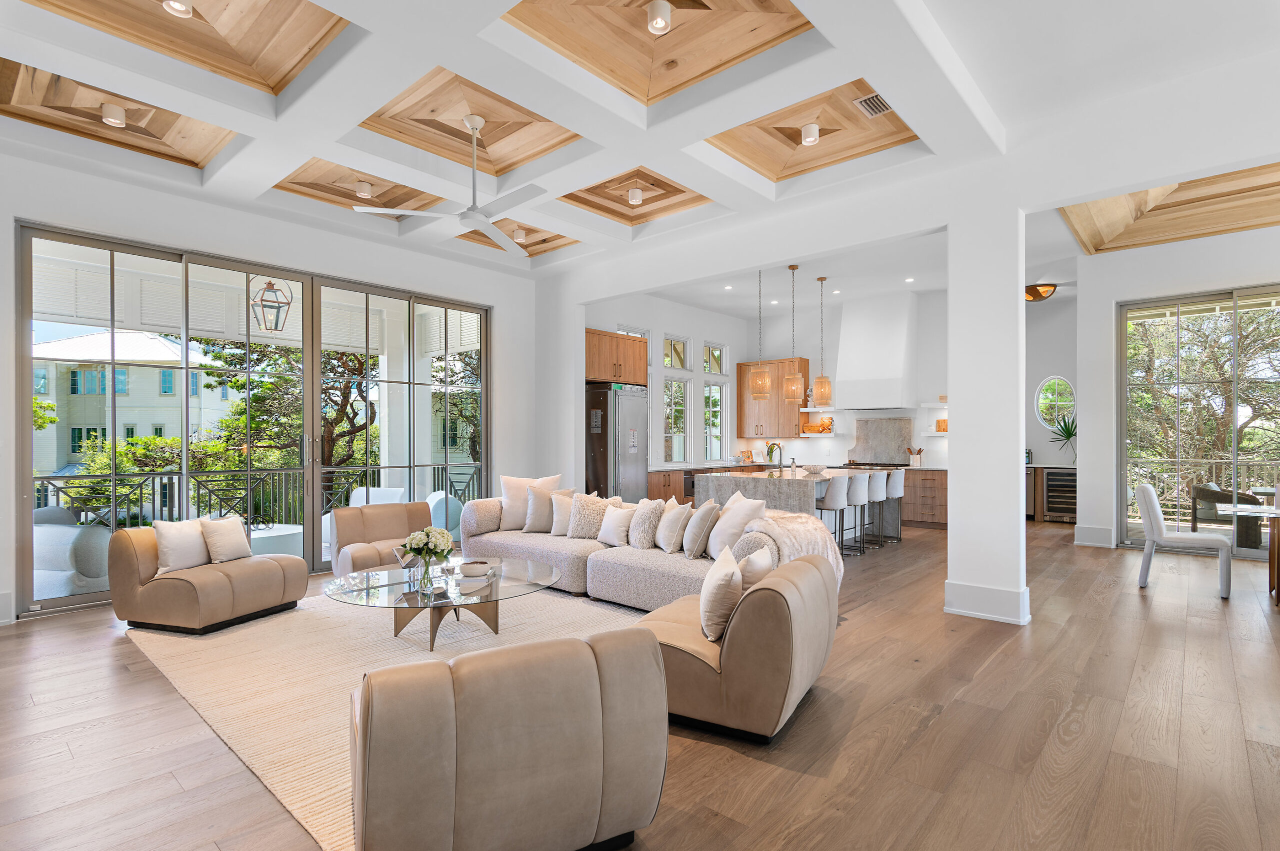 Spacious, modern open-concept living room and kitchen with beige sofas, wood accents, large windows, coffered ceiling, and neutral decor—perfect for entertaining your a-list guests. Dining area is visible to the right through wide doorways.