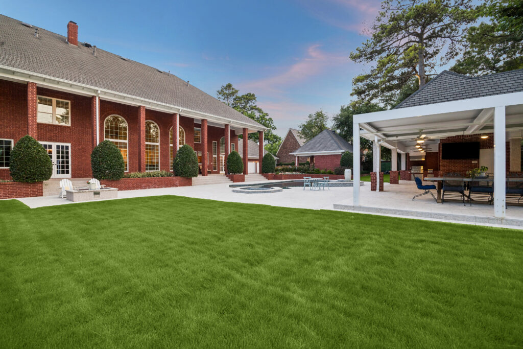 A large brick house with tall windows opens to a spacious backyard featuring a green lawn, patio seating, a covered outdoor dining space, and a small pool, all surrounded by tall trees at sunset.