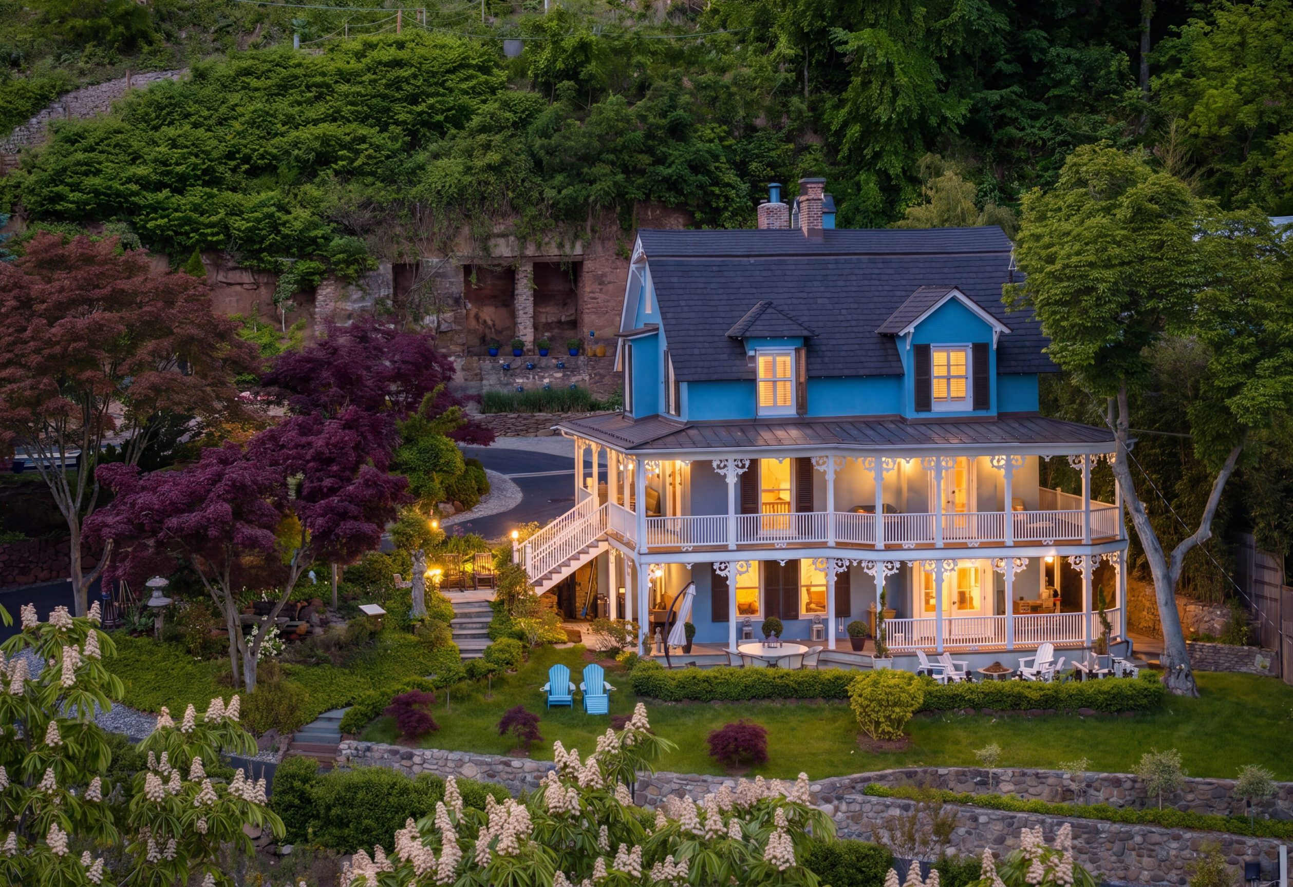 A charming blue and white two-story house with a wraparound porch is lit warmly in the evening, surrounded by lush trees and a green lawn. Ideal for an A-list gathering, June 2026, with Adirondack chairs and blooming bushes completing the scene.