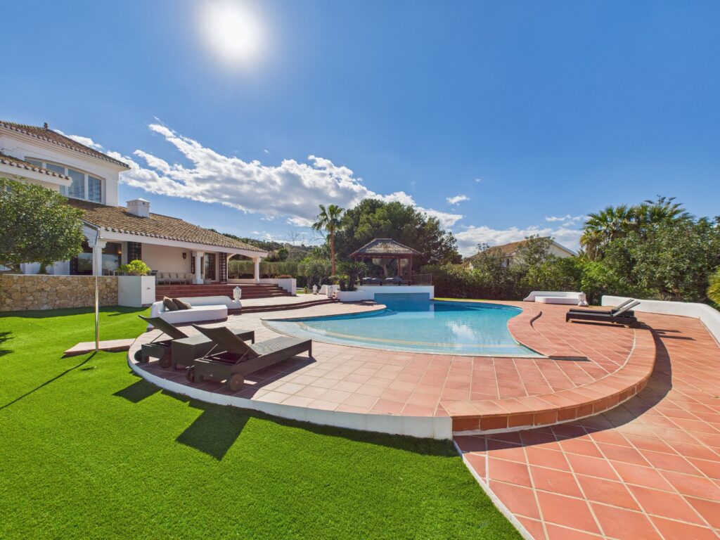 A sunny backyard scene features a large swimming pool surrounded by terracotta tiles, lounge chairs, green artificial grass, and a modern house in the background under a clear blue sky.