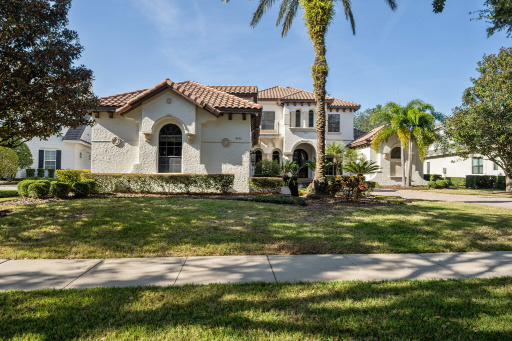Two-story Mediterranean-style house with white stucco walls, terracotta roof tiles, and arched windows, surrounded by palm trees and vibrant landscaping that glows under the comforting light of cancer season’s sunny days.