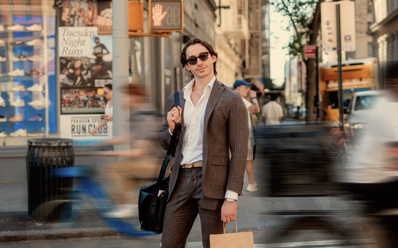 A man in a suit and sunglasses stands on a city street corner, holding a brown paper bag and a black shoulder bag. People and cyclists blur past him, capturing the busy urban environment.