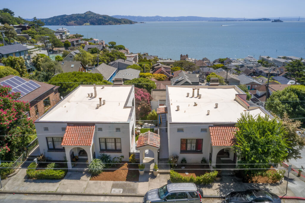 Aerial view of two white stucco houses with red tile roofs on a coastal street, surrounded by greenery and neighboring homes, overlooking a bay and distant hills under the clear sky.