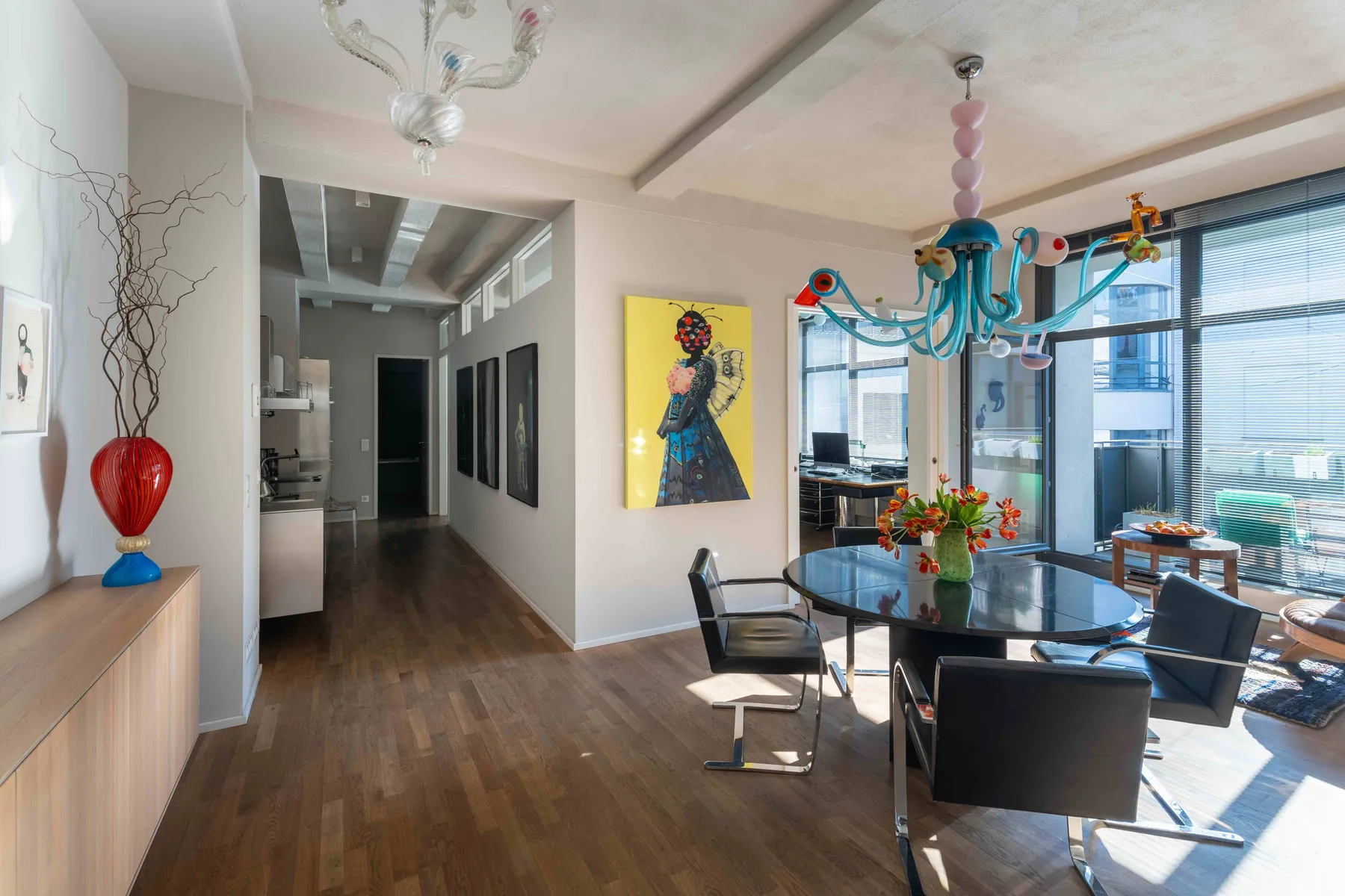 A modern dining room with a round glass table, four black chairs, and vibrant artwork feels ready for an A-list June 2026 dinner party. Large windows with blinds and a colorful chandelier let natural light fill the space.