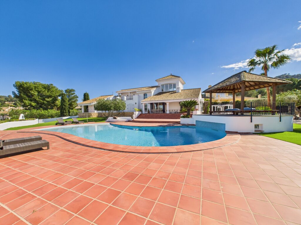 A large, modern villa with a tiled patio, infinity pool, sun loungers, and a wooden gazebo, surrounded by palm trees and greenery—perfect for relaxing under the clear blue sky.