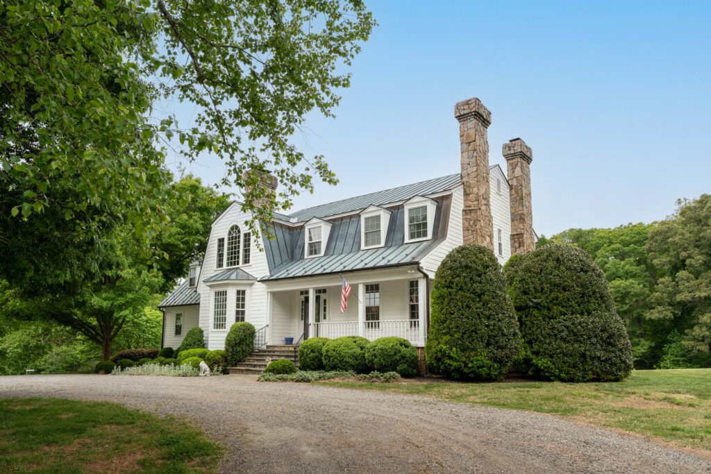 A large white house with a metal roof, two stone chimneys, and dormer windows sits near green trees. During cancer season, an American flag hangs by the entrance, and a gravel driveway curves in front of trimmed bushes and landscaping.