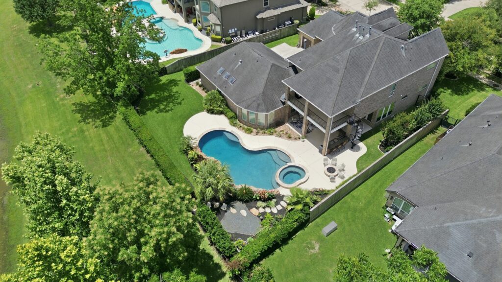 Aerial view of a suburban backyard, featuring an irregularly shaped swimming pool, spa, patio area, landscaped garden, and green lawn, all surrounded by a wooden fence and neighboring houses.
