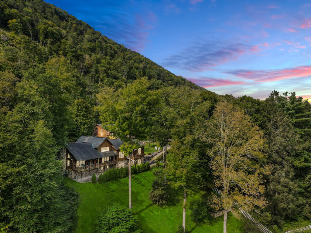 A rustic wooden house is nestled among lush green trees at the base of a forested hillside.