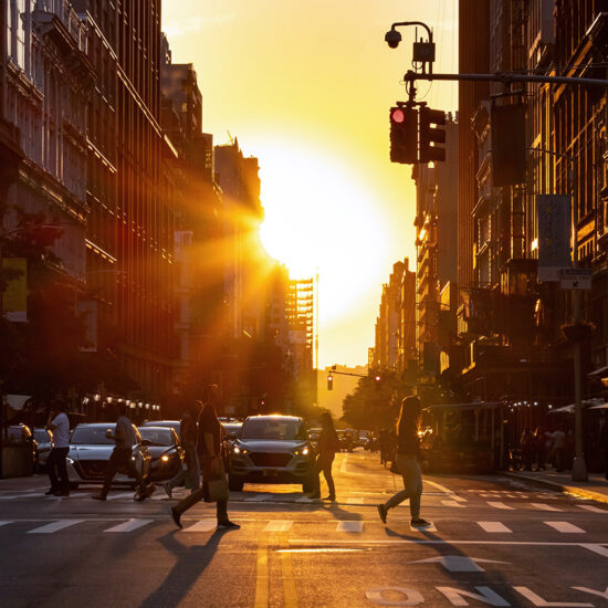 A city street at sunset with people crossing the road, cars stopped at a traffic light, and tall buildings lining both sides—capturing the vibrant life and energy of the Manhattan real estate market bathed in warm golden sunlight.