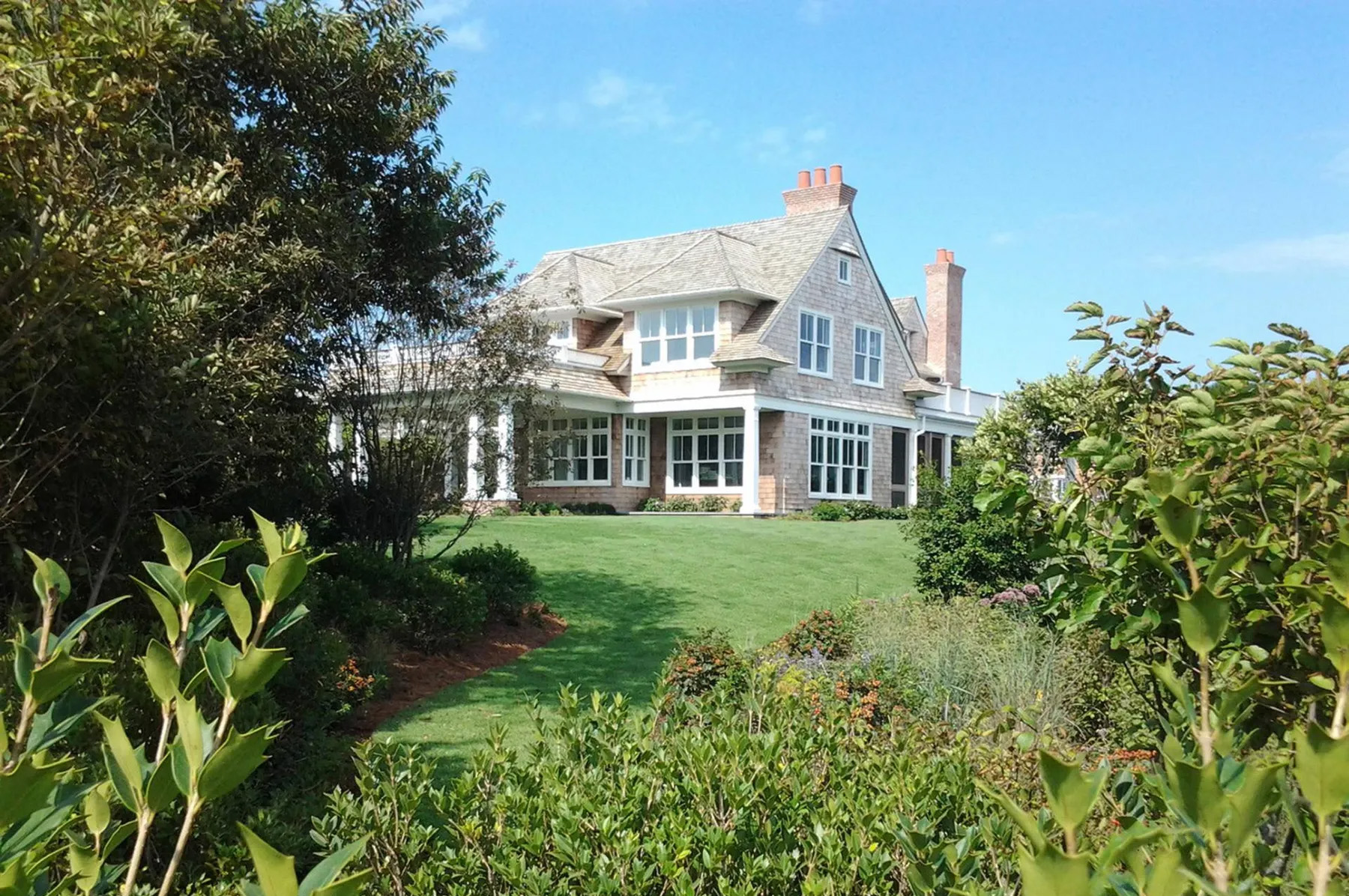 A large, elegant house with many windows and a stone chimney sits amid lush green gardens and trees under a clear blue sky—perfect for hosting your A-list June 2026 gatherings.